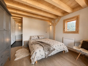 Een slaapkamer met een houten plafond, een bed met behang, een zacht vloerkleed, een stoel met een vacht en een raam met uitzicht op bomen.