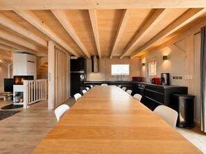 Ein geräumiger Raum mit einem langen Holztisch und weißen Stühlen, einer Küche mit modernen Geräten sowie einem Kamin.