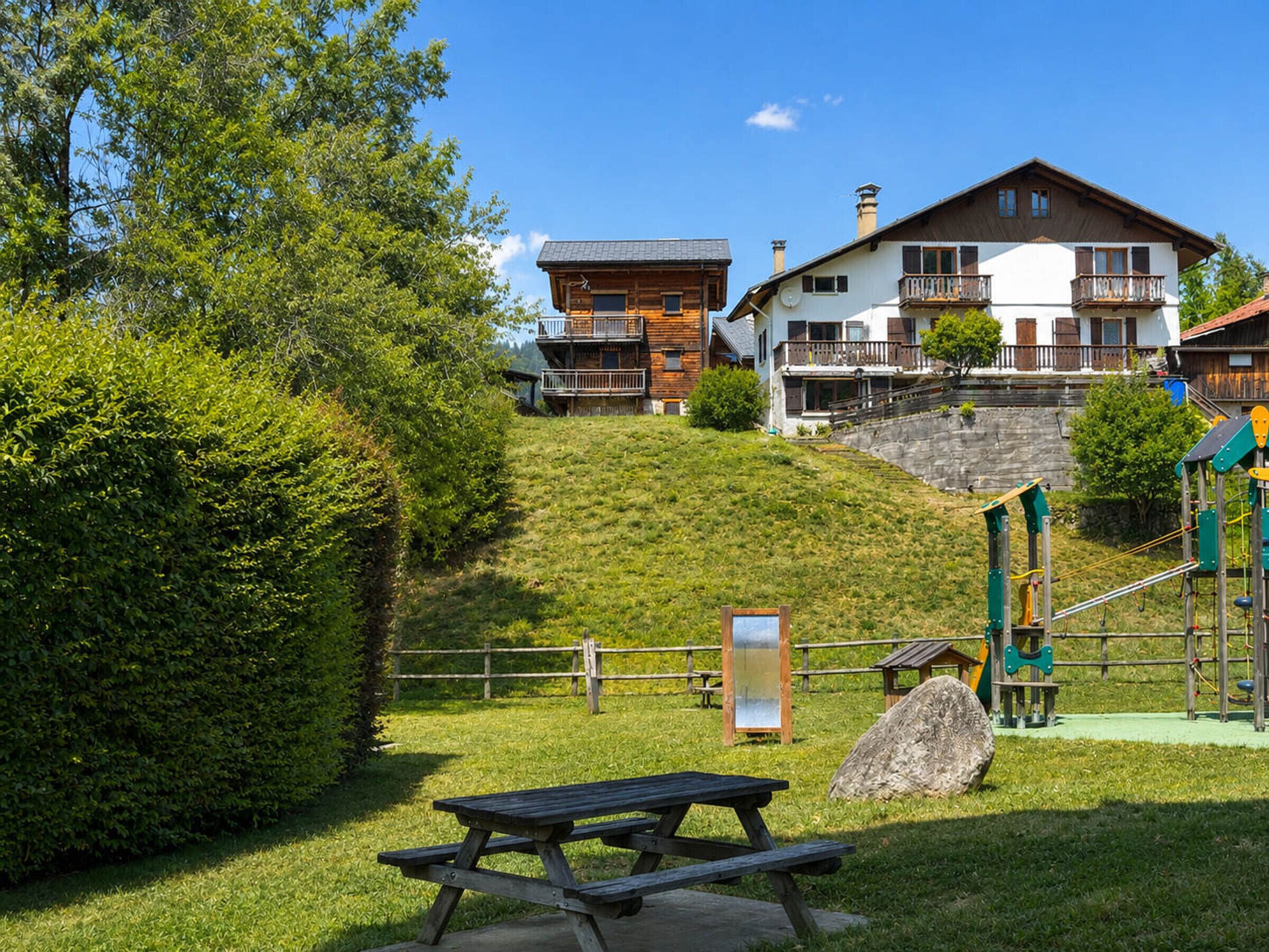 En ejendom på en skråning med en træhytte, en hvid bygning med balkoner og en legeplads.