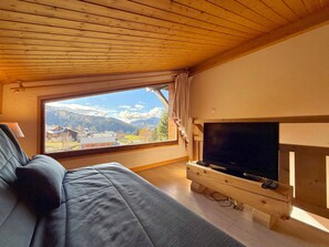 Une pièce avec un plafond en bois, un canapé, un téléviseur posé sur un meuble en bois et une fenêtre donnant sur la montagne.