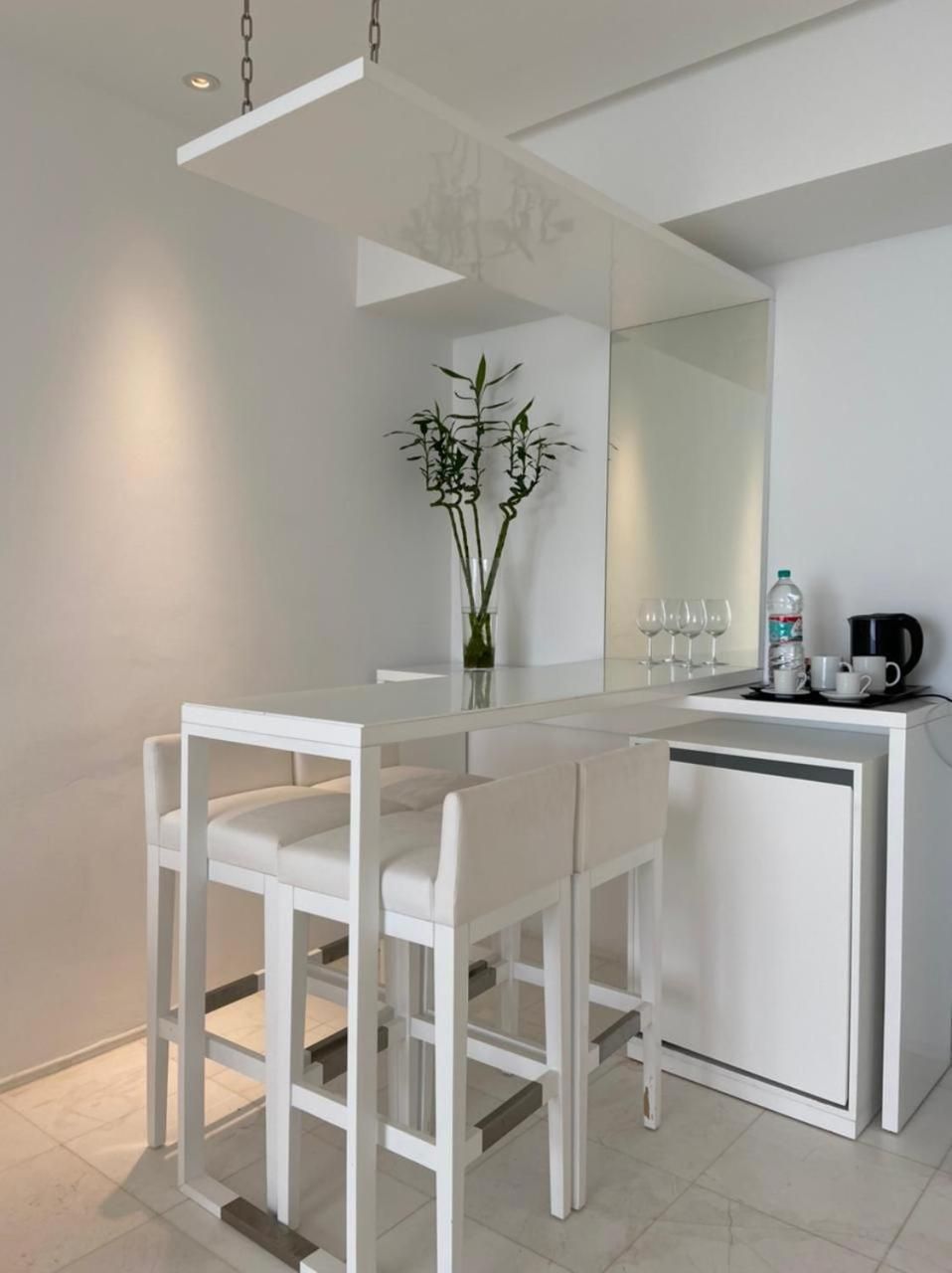 Una habitación moderna y minimalista con una mesa y sillas blancas para comer, un mueble blanco pequeño, un espejo y una planta en maceta.