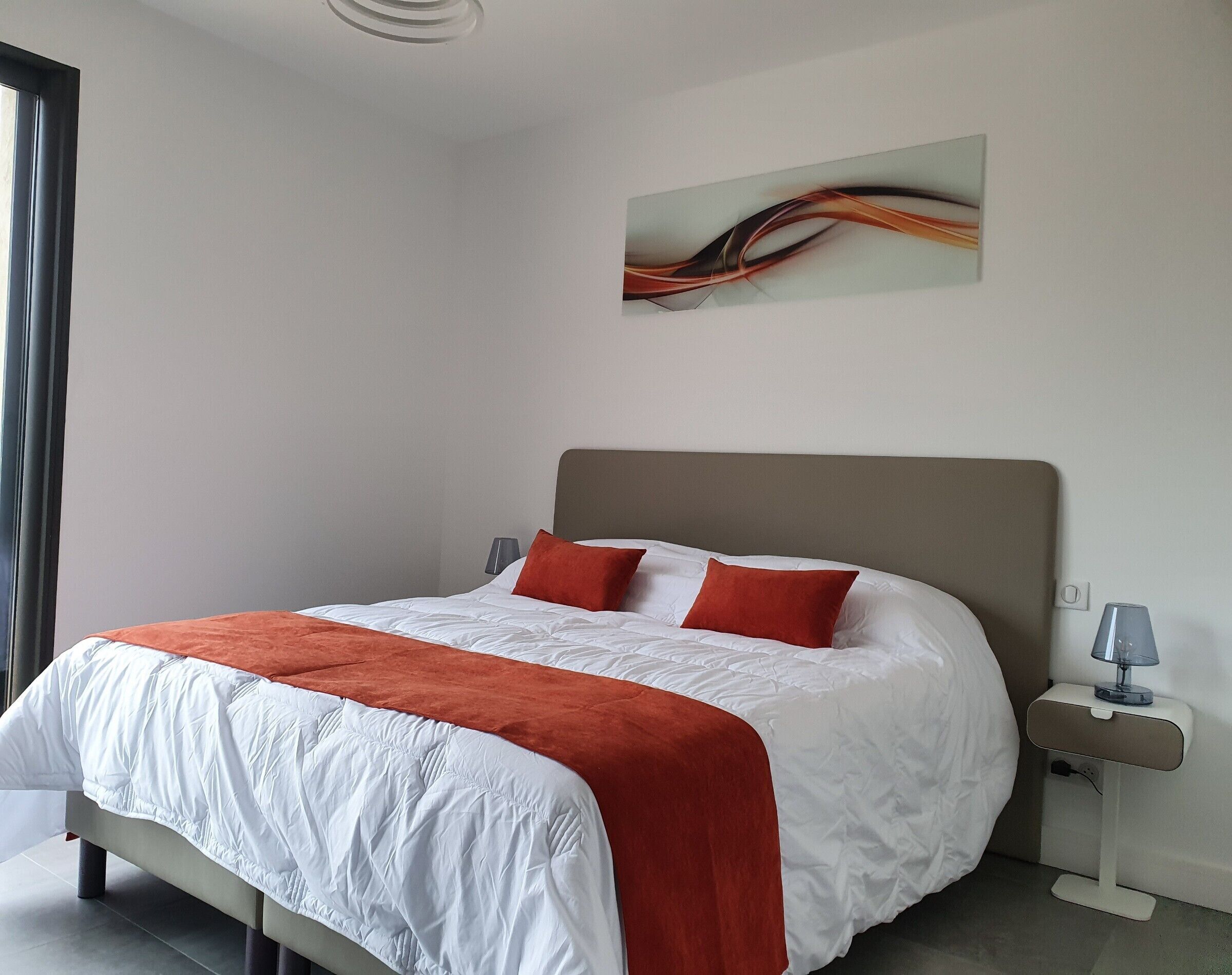 Een net opgemaakt bed met een wit-oranje dekbedovertrek, een grijs hoofdbord en twee rode kussens. Aan de rechterkant staat een moderne lamp op een nachtkastje.