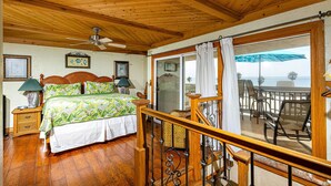 Une chambre avec un plafond en bois, un lit recouvert d’une couvre-lit à motifs floraux, une table de chevet avec une lampe, et une vue sur l’océan par une porte coulissante.