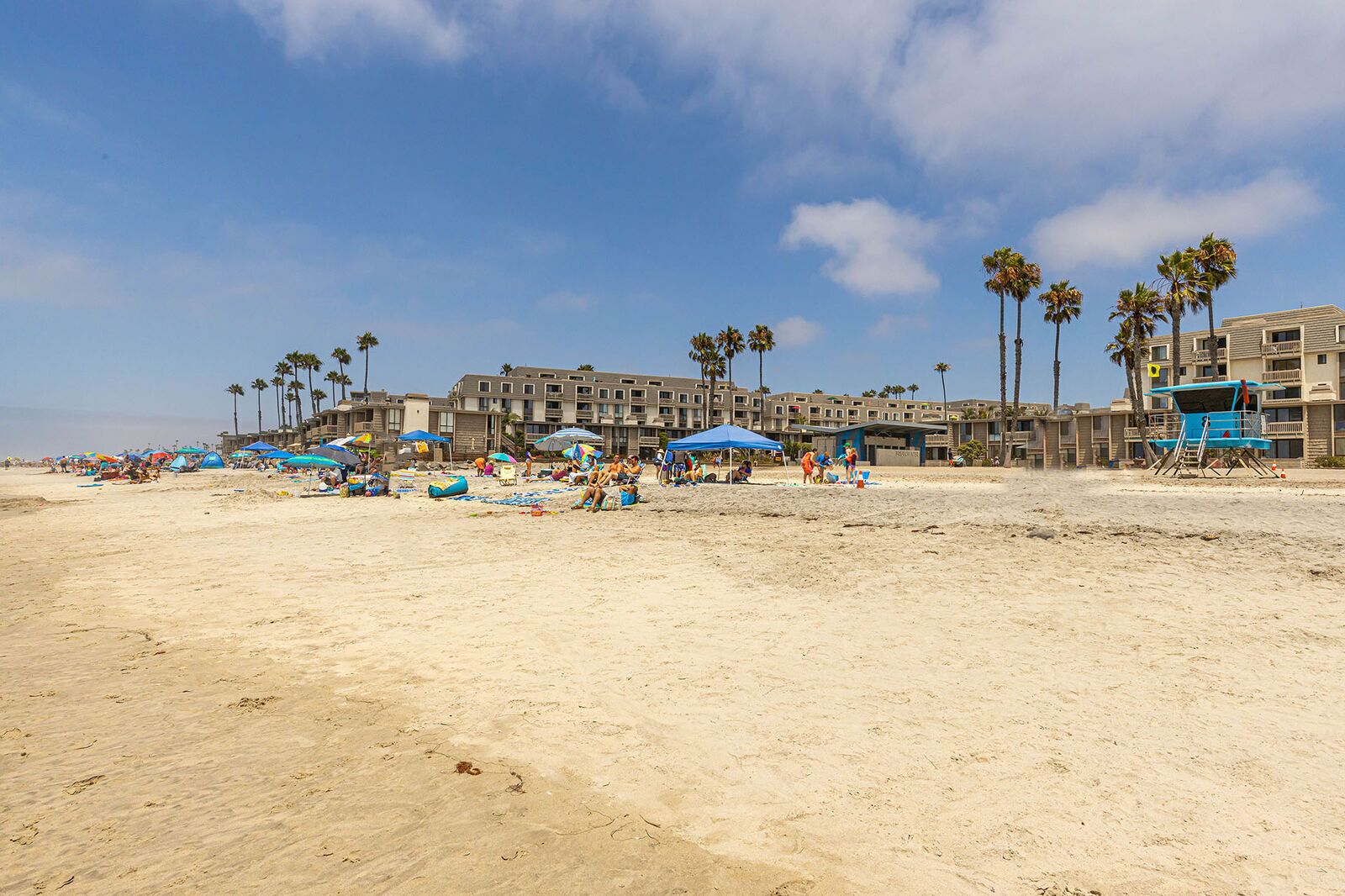 Une plage avec de nombreux parasols et des chaises de plage, des palmiers et, au fond, un bâtiment doté de balcons.