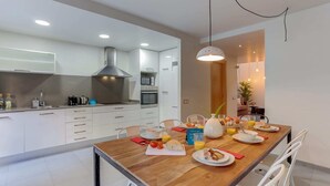Eine moderne Küche mit weißen Schränken, einem Holztisch und einer Frühstücksecke mit Tellern, Gläsern und einer Obstschale.