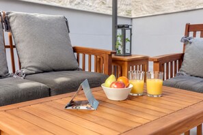 A wooden table with a bowl of fruit and two glasses of orange juice, a folded magazine, and a small plant on a balcony with wooden chairs and cushions.