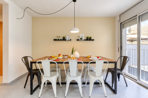 A dining area with a wooden table, white chairs, a hanging light fixture, and a view through a glass door.