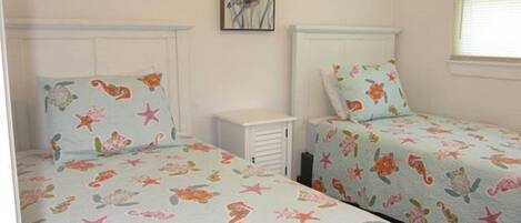 Une chambre avec deux lits, chacun recouvert d’une courtepointe aux motifs floraux et marins. Une petite table de chevet blanche est placée entre les deux lits, et un tableau encadré est accroché au mur au-dessus du lit situé à gauche.