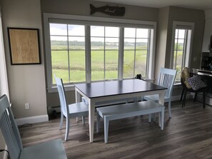 A dining area with a large window offering a view of a grassy field. There is a framed map on the wall and a fish-shaped sign above the window. - Modern house at Wells Beach - Available Winter Rentals Off Season-reduce rate. (Wells)