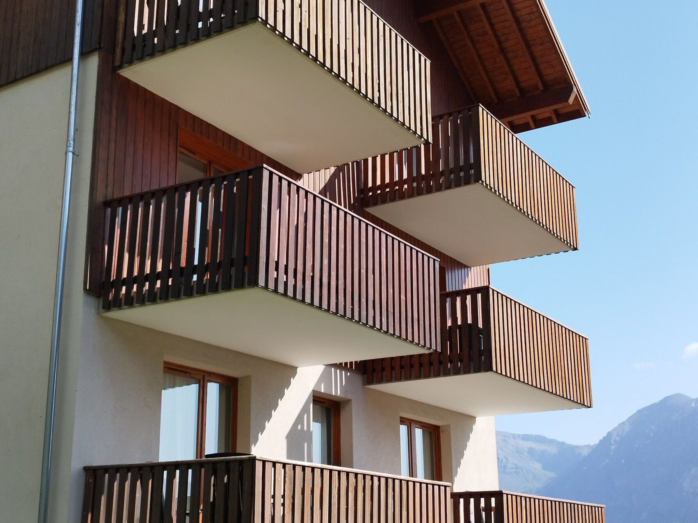 Un edifici de diverses plantes amb balcons, grans finestres i un paisatge de muntanyes al fons.