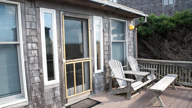 Eine Holzterrasse mit zwei Adirondack-Stühlen, einem Picknicktisch, einer Glasbiflügeltür und einem kleinen Veranda-Bereich mit Fußmatte.
