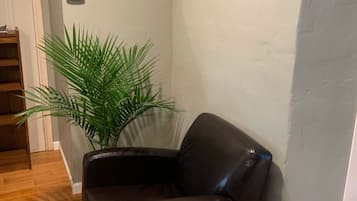 A corner room with a leather armchair, a large potted plant, a framed picture on the wall, and a thermostat.