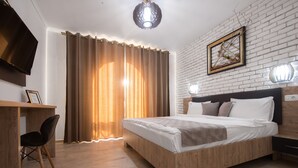 Ein Hotelzimmer mit einem hölzernen Bett, einem Schreibtisch mit Stuhl, einem Fernseher und einem großen Fenster mit Vorhängen.