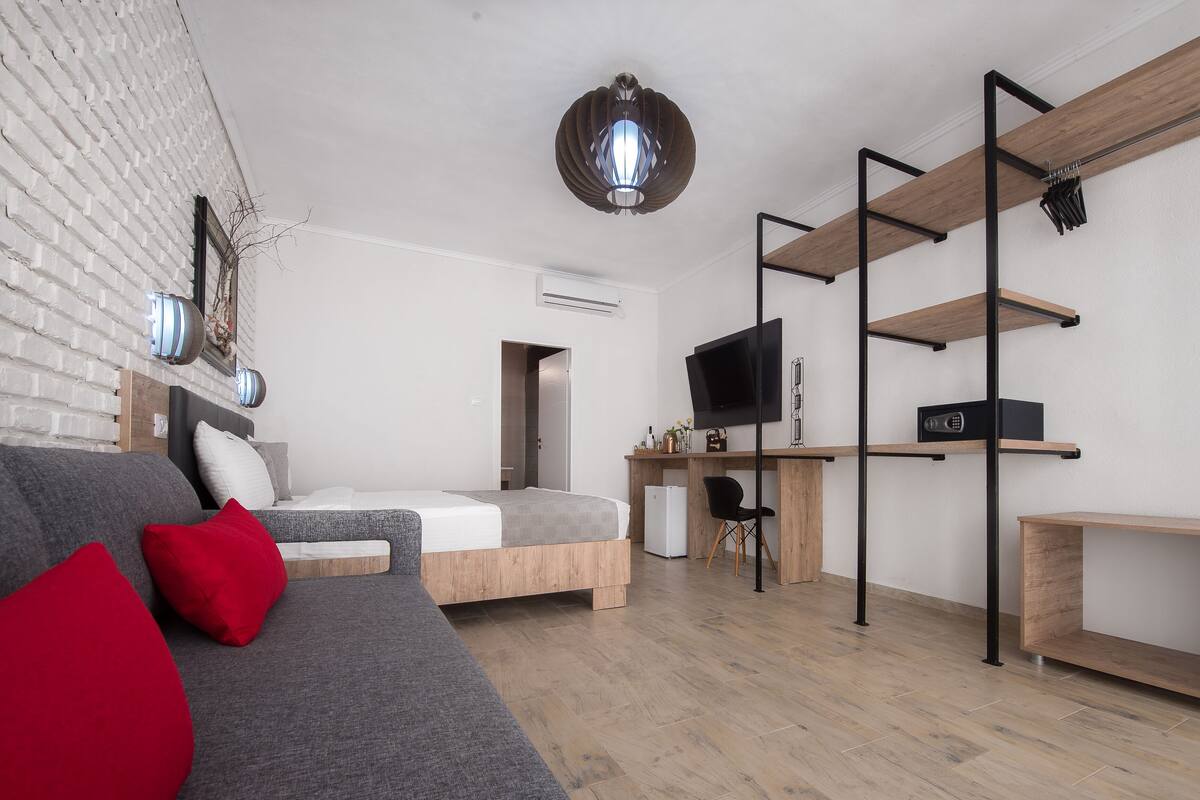 A modern, well-lit bedroom with a bed, a grey sofa with red cushions, a TV, and a desk with shelves.