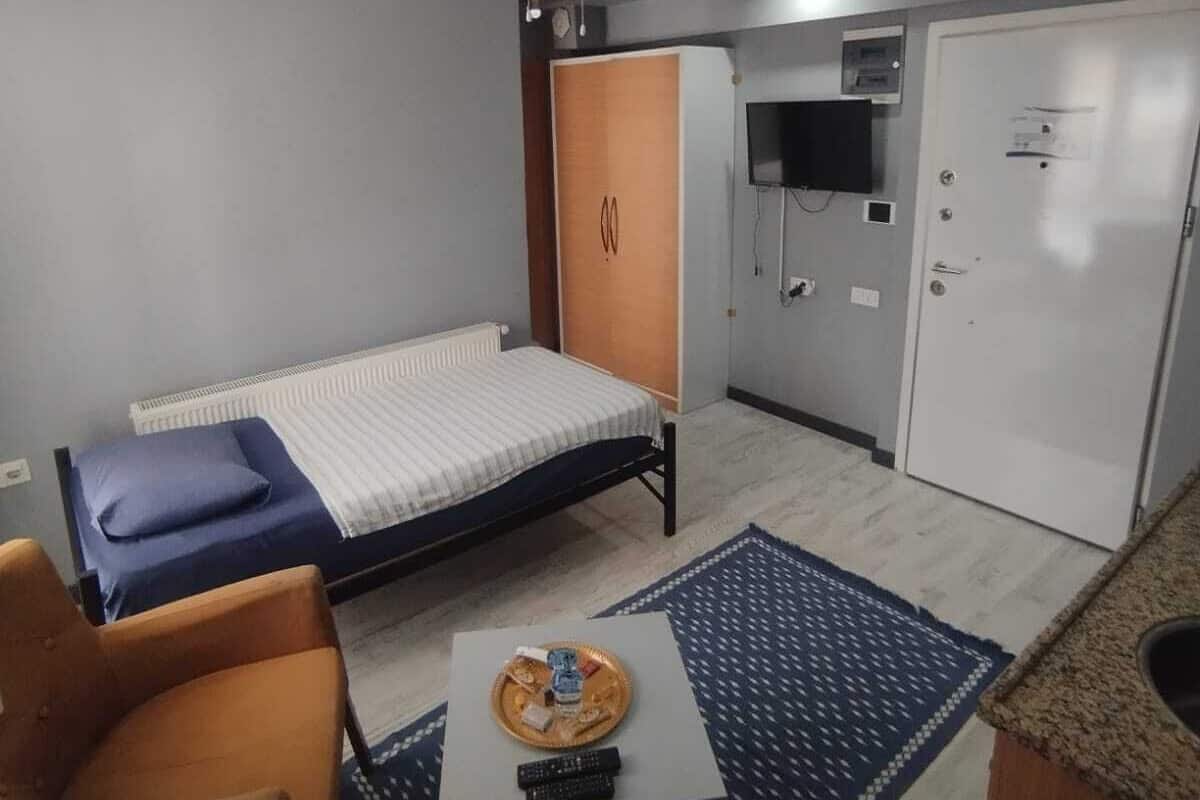 Ein kleines Hotelzimmer mit einem Einzelbett, einem Tisch mit Fernbedienung, einem Sessel, einem Kleiderschrank und einem Deckenventilator.