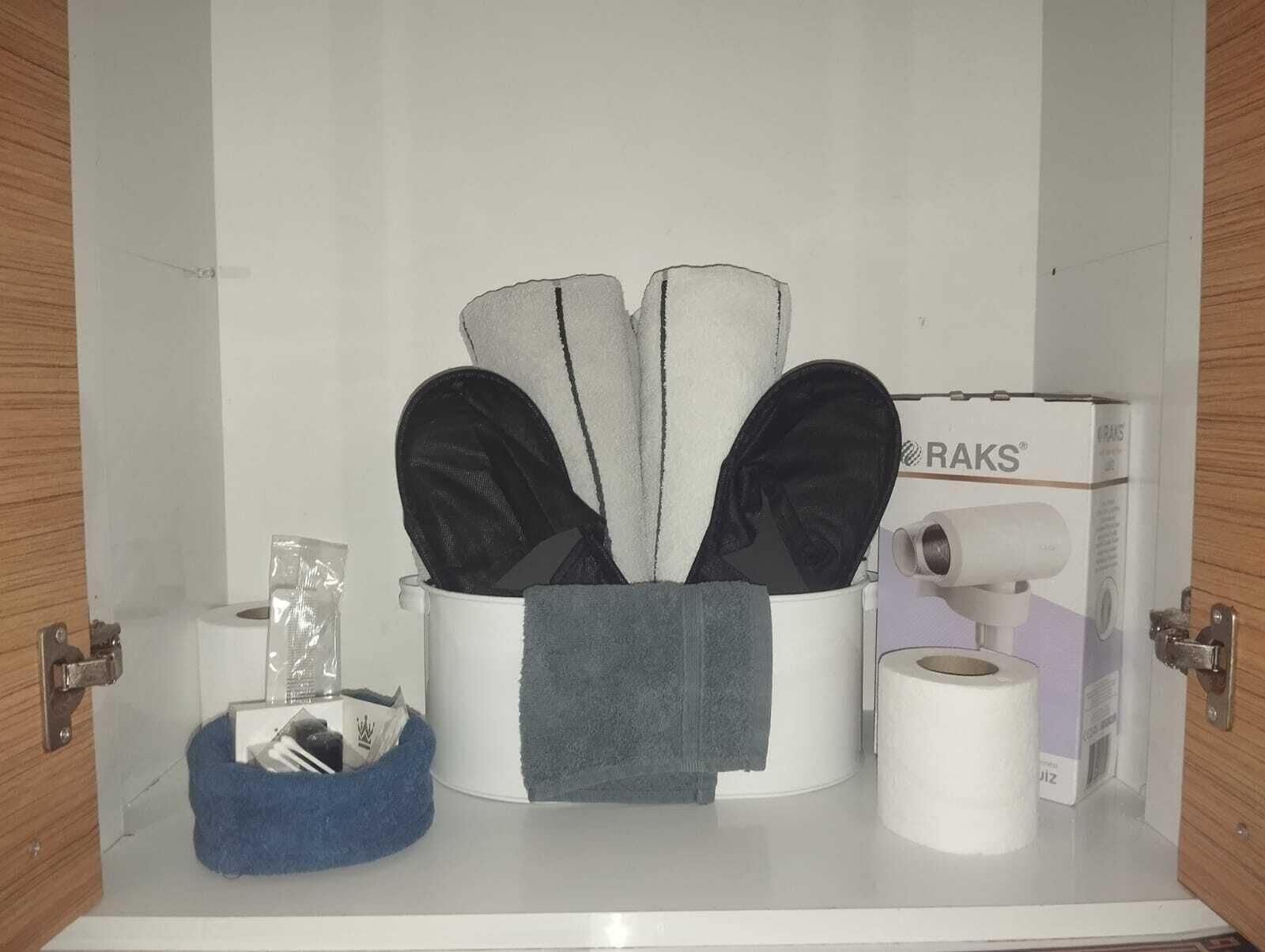 Een opslagruimte met een blauwe mand, een witte bak met zwarte spullen, en een doos met de tekst 'RAKS'.