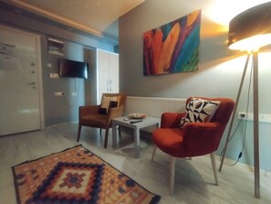 Een moderne woonkamer met een bank, fauteuil en een salontafel. Op de muur hangt een kleurrijk schilderij en er staat een vloerlamp.