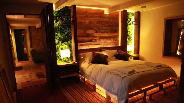 A bedroom with a wooden headboard, a large bed with pillows, a bedside table with a lamp, and a view of greenery through an open door. - The Lodge Rooms (Coleford)