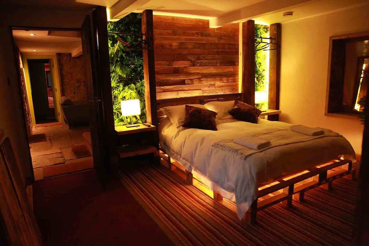Ein Schlafzimmer mit Holz-Kopfteil, einem großen Bett mit Kissen, einem Nachttisch mit Lampe und Blick auf Grün durch eine offene Tür.