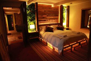 A bedroom with a wooden headboard, a large bed with pillows, a bedside table with a lamp, and a view of greenery through an open door. - The Lodge Rooms (Coleford)