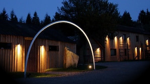 A rustic wooden building with a large archway entrance, illuminated by exterior lights, surrounded by trees. - The Lodge Rooms (Coleford)