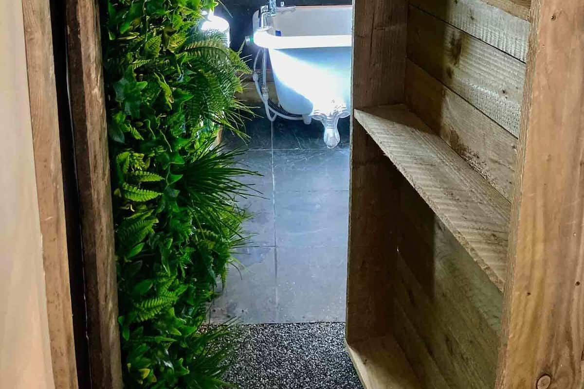 Ein schmaler Flur mit einem Holzregal rechts und einer Wand aus Grünpflanzen links, der zu einem Badezimmer mit Badewanne und Waschbecken führt.