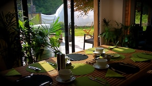 A dining table set with green placemats, white cups, and cutlery, overlooking a garden with a white tent and potted plants. - The Lodge Rooms (Coleford)