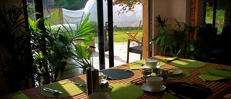 A dining table set with green placemats, white cups, and cutlery, overlooking a garden with a white tent and potted plants.