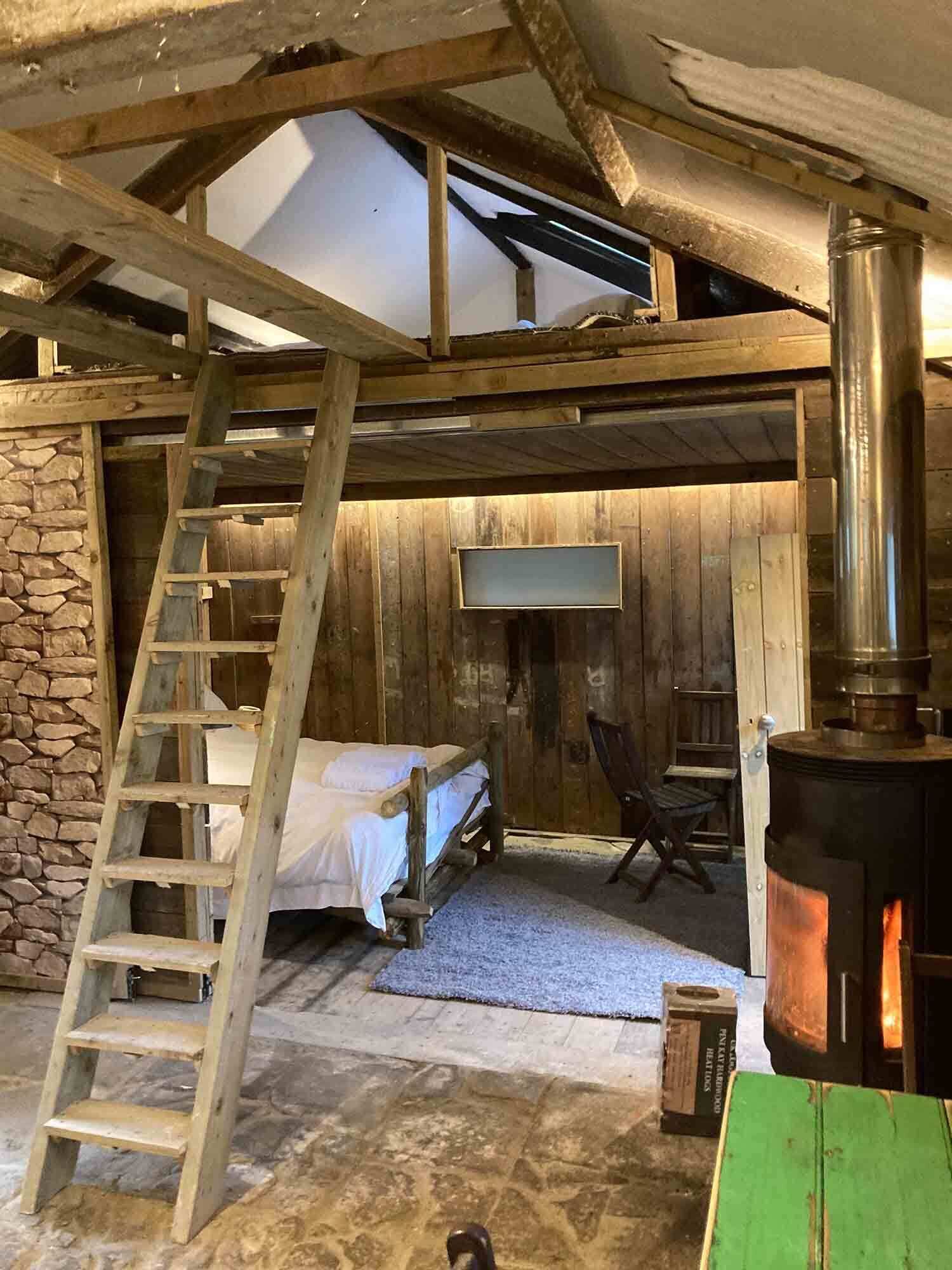 A rustic room with a stone wall, wooden beams, a bed, a ladder, a fireplace, and a television.