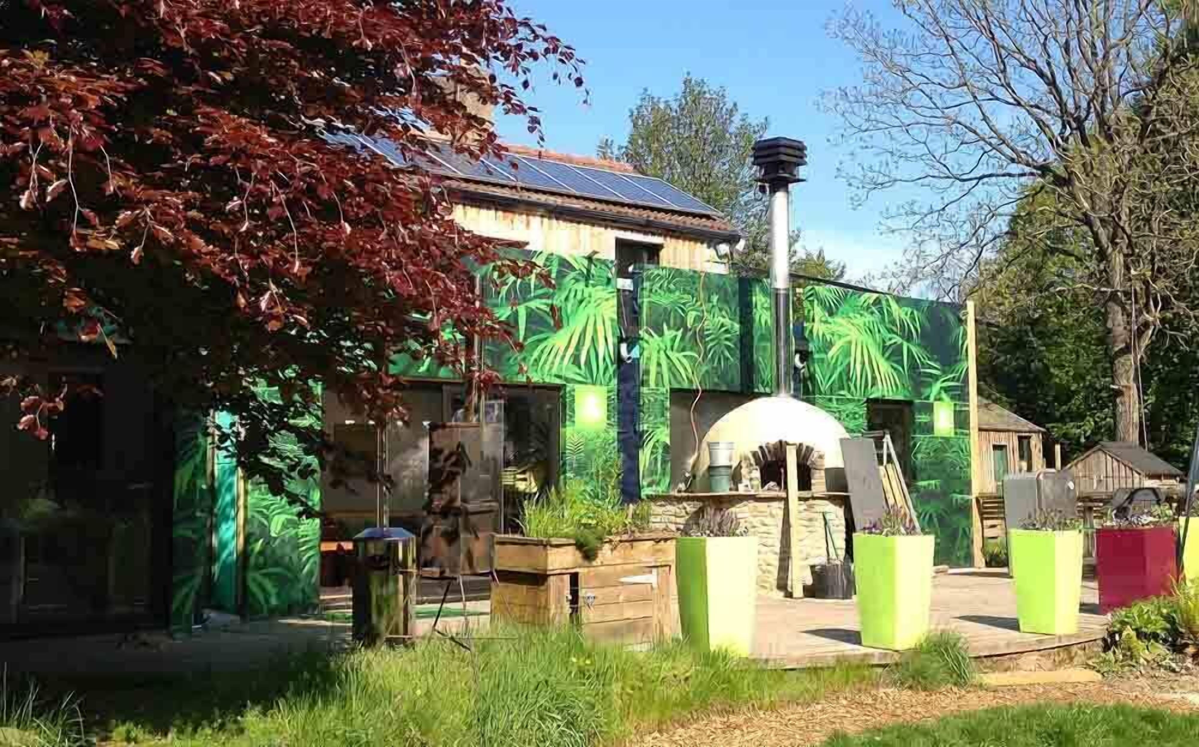 A rustic building with a solar panel roof, surrounded by lush greenery and a stone structure in the center.