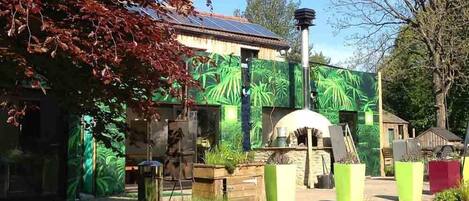 Un bâtiment rustique surmonté d’un toit équipé de panneaux solaires, entouré d’une végétation luxuriante et agrémenté d’une structure en pierre au centre.