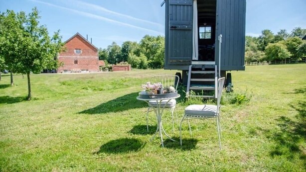 Uma pequena construção aberta com uma mesa e cadeiras na frente, situada num campo gramado com um edifício de tijolos ao fundo.