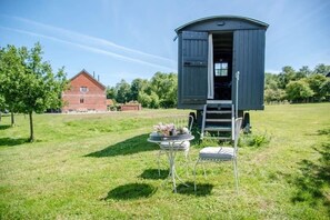 A small, open-sided structure with a table and chairs in front, set in a grassy field with a brick building in the background. - Abbey Shepherds Hut, Eye (Air Manage Suffolk) (Eye)
