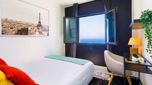 Ein Hotelzimmer mit einem Bett, einem Schreibtisch mit Lampe, einem Stuhl, einem Fenster mit Vorhängen und einem Stadtbildgemälde.