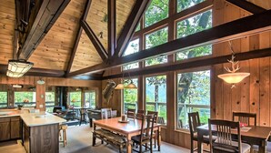 A spacious dining area with wooden tables and chairs, large windows offering views of the outdoors, and a high ceiling with exposed beams.