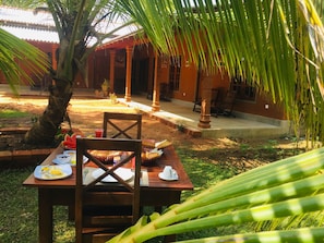 An outdoor dining area with a table set for two, surrounded by palm trees and a building with a tiled roof.