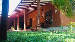 A building with a covered porch, columns, and a garden area with grass and plants. - Jaye Eco Lodge (Habarana)