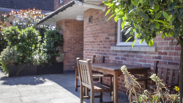 A brick patio with a wooden table and chairs, surrounded by greenery and a brick wall.