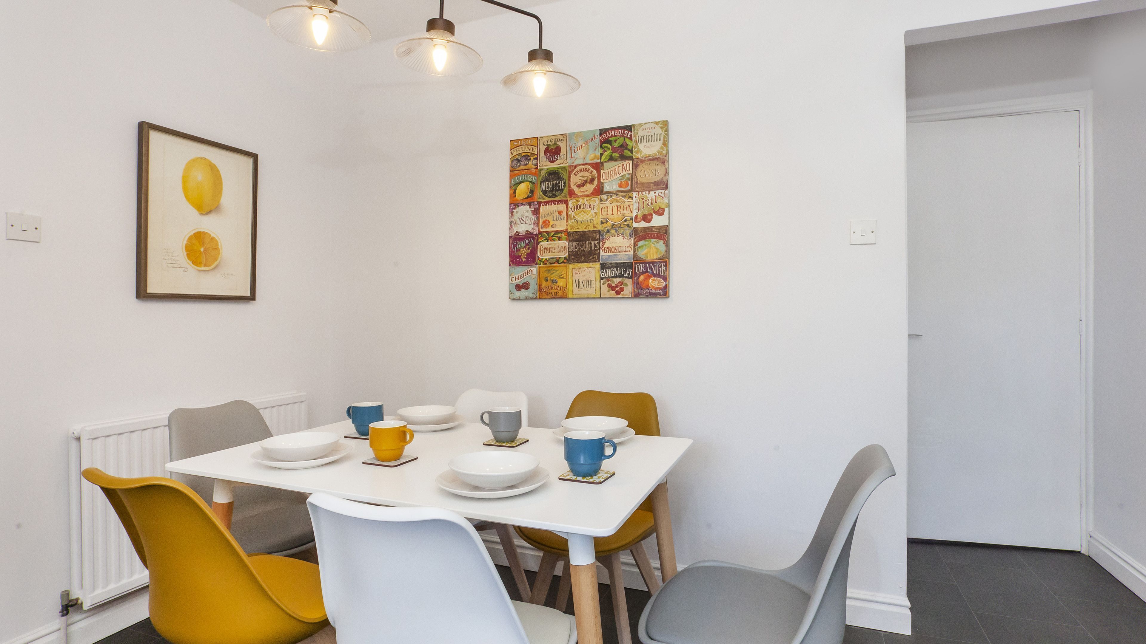 A dining area with a white table, yellow and gray chairs, and a colorful fruit-themed wall art.