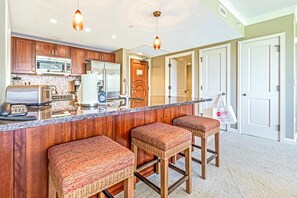 A kitchen with wooden cabinets, a granite countertop, and three wicker bar stools. - Honua Kai | Ocean View 2 BR | Free Car w/ 6+ Nights | HKH-348 (Lahaina)