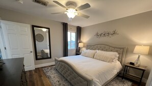Ein Schlafzimmer mit einem karottenbesetzten Kopfteil, einem großen Spiegel, einem Deckenventilator, einem Nachttisch mit Lampe und einem gemusterten Teppich.