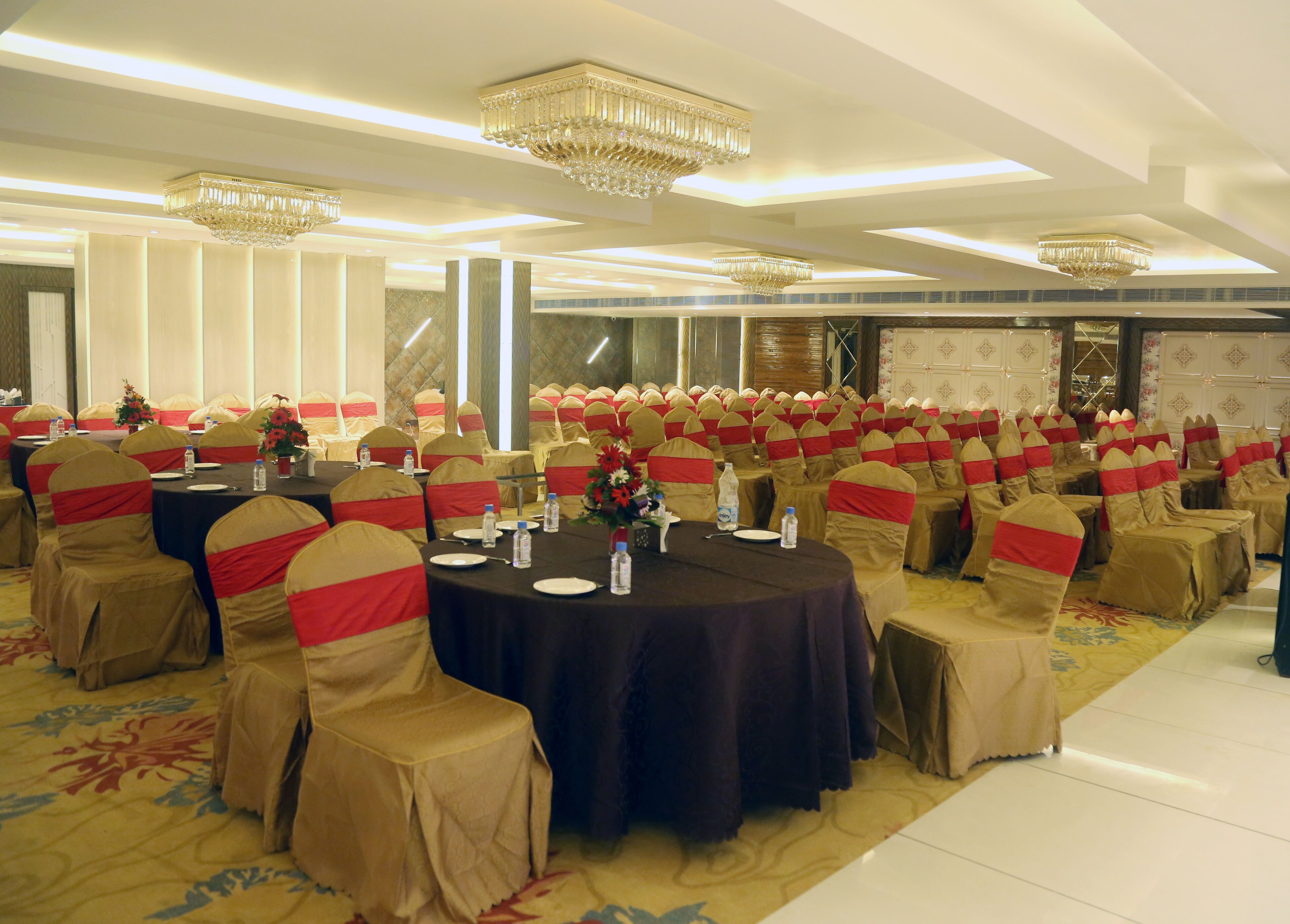 A banquet hall with round tables, chairs with red sashes, and chandeliers.