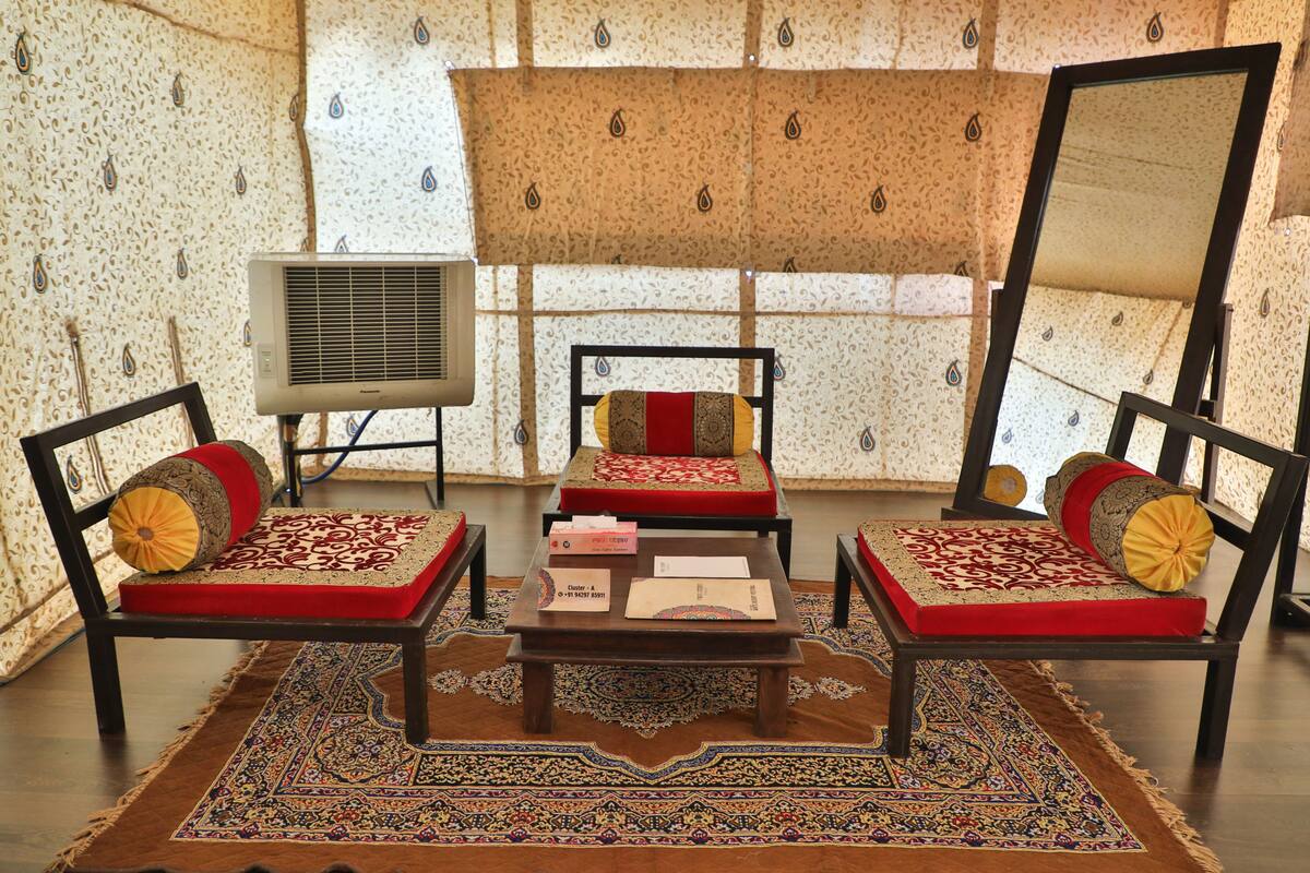 A room with a traditional design, featuring a sofa set with red cushions, a matching ottoman, and a patterned rug. There is a small wooden table with a book and a box, and a wall-mounted air conditioner.