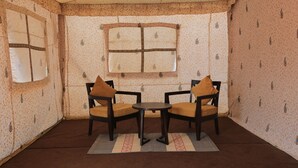 A room with two wooden chairs and a small table, patterned wallpaper, and a window with a cross design. - Rann Utsav The Tent City (Bhuj)
