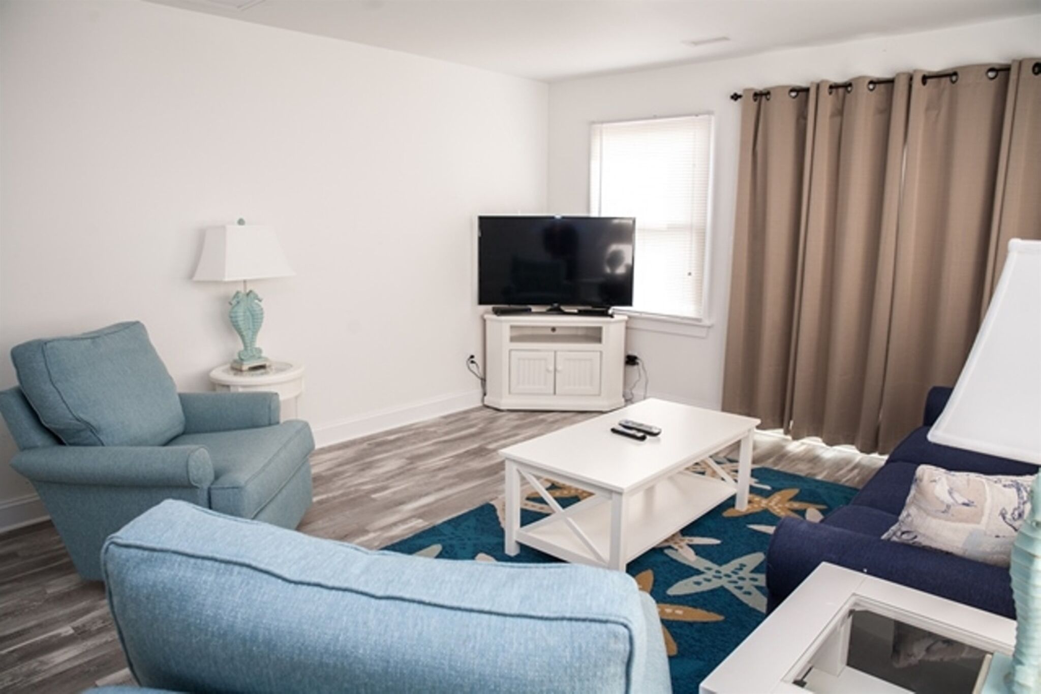 A modern living room with a blue armchair, a white coffee table, a flat-screen TV on a white cabinet, a window with beige curtains, and a blue patterned rug.