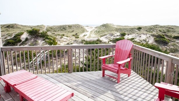 Une terrasse en bois avec des fauteuils Adirondack rouges et un banc, donnant sur une plage bordée de dunes et sur un sentier.