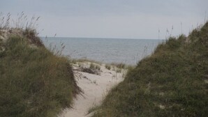 Un étroit sentier sablonneux qui mène à une plage bordée de dunes herbeuses de chaque côté.