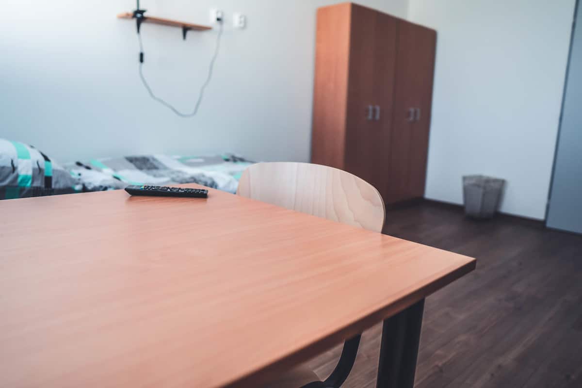 A room with a wooden table, a bed with a blanket, a wardrobe, and a trash bin.