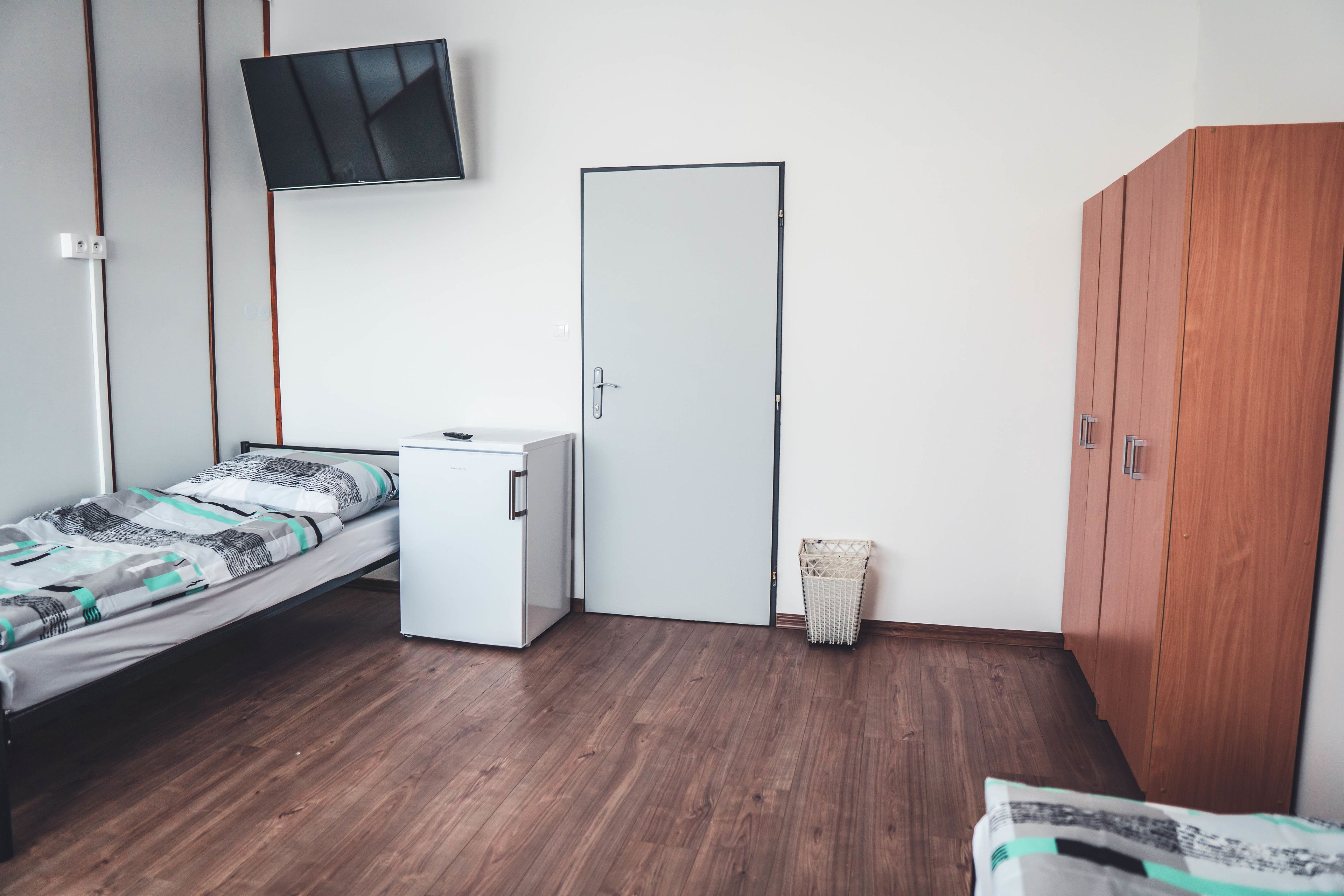 A room with a bed, a small white cabinet, a wooden wardrobe, a door, and a wall-mounted TV.