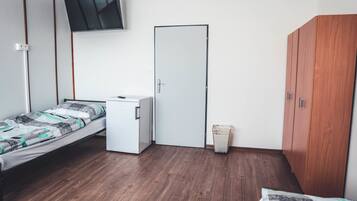 A room with a bed, a small white cabinet, a wooden wardrobe, a door, and a wall-mounted TV.
